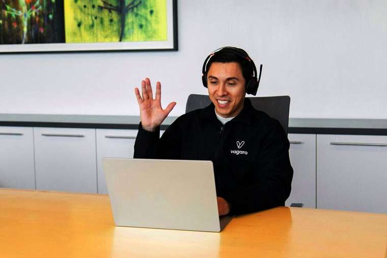Hombre joven con auriculares saludando frente a una notebook
