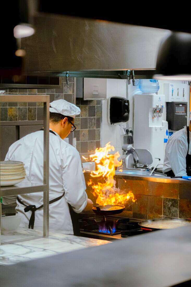 Cocinero con chaquetilla blanca salteando en una sartén con llamas