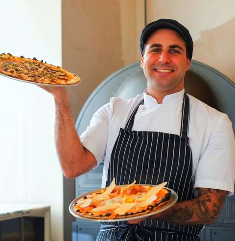 Pizzero sonriendo con delantal a rayas y una pizza en cada mano