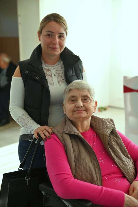 Cuidadora de pie junto a una mujer mayor en silla de ruedas, las dos mirando a cámara