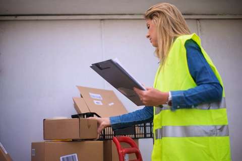 Empleada de depósito con chaleco reflectivo revisando una planilla junto a unas cajas