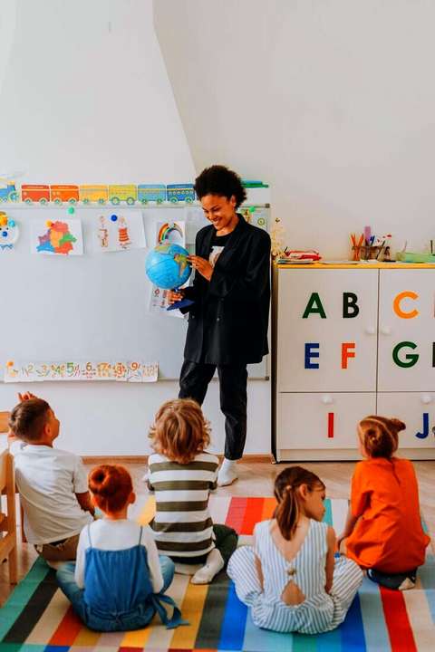 Docente con un globo terráqueo frente a niños sentados en el piso, vistos de espaldas