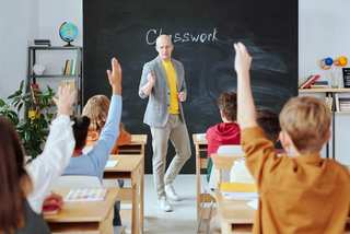 Docente de pie frente al pizarrón con alumnos levantando la mano