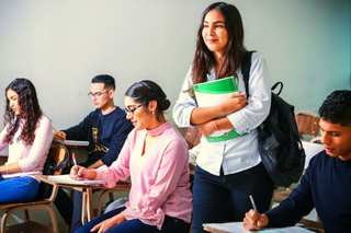 Estudiante con mochila y carpeta sonriendo en un aula con compañeros