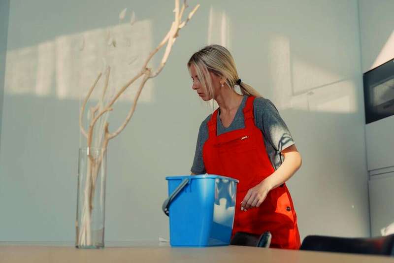 Mujer con delantal naranja y un balde celeste limpiando en una cocina
