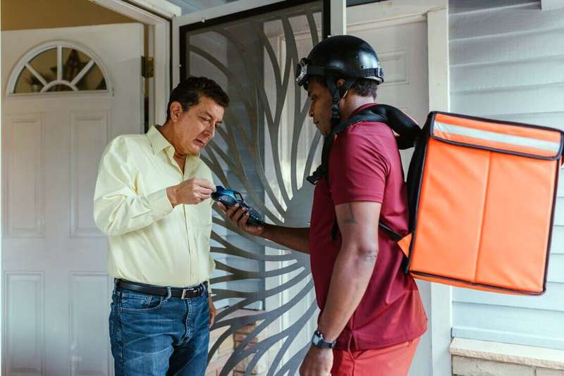 Repartidor con casco y mochila térmica entregando un pedido a un cliente en la puerta de su casa