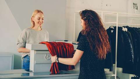 Vendedora sonriendo detrás del mostrador mientras una clienta le muestra una prenda roja en un local de ropa